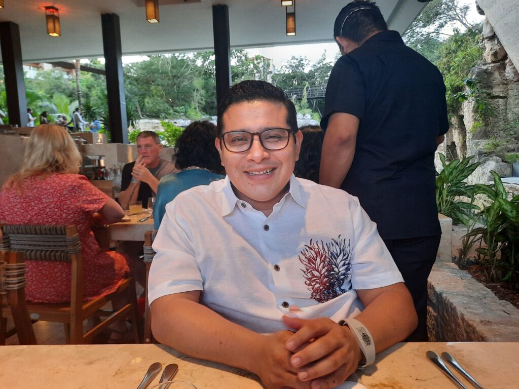 Hombre sonriente en un restaurante con un fondo de vegetación y otras personas comiendo.